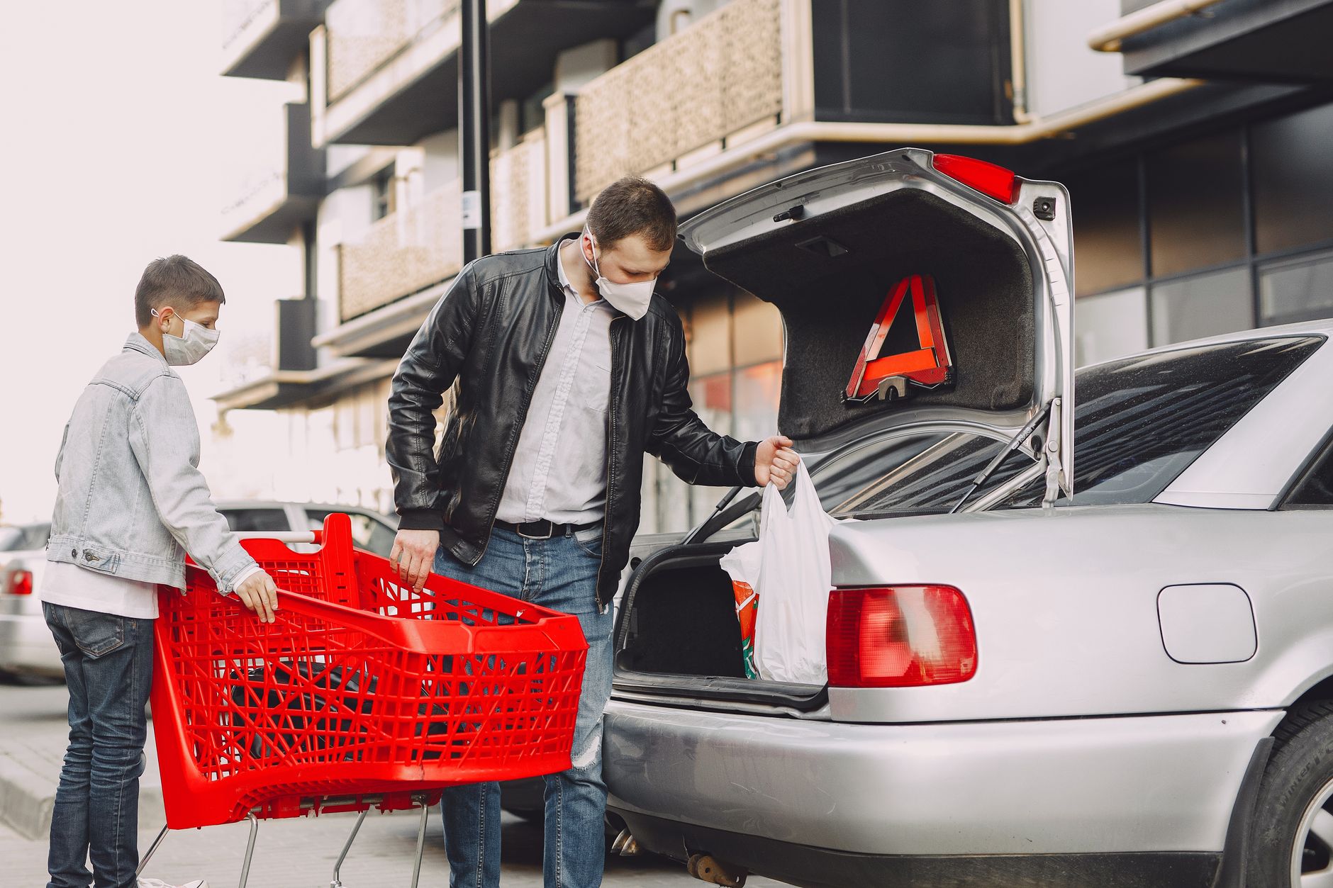 Ojciec i syn w maskach pakują zakupy do bagażnika na parkingu